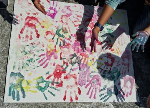 A colorful canvas featuring a collage of handprints in various shades, including red, pink, green, yellow, and blue. The handprints are overlapping and scattered across the surface, with some hands still applying paint.