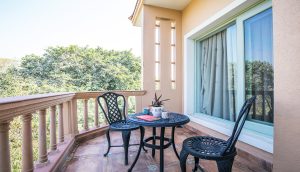 This photograph shows a calm, sunlit balcony designed for quiet relaxation. A small round black metal table sits at the center, paired with two matching chairs featuring decorative cutout patterns. On the table are a potted plant, patterned mugs, and a book, suggesting a peaceful moment for reading or enjoying tea. The balcony floor is tiled in war