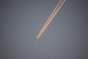 A passenger plane high in a clear blue sky as seen from below, with its twin contrails extending out of frame and golden in color in the sunlight.