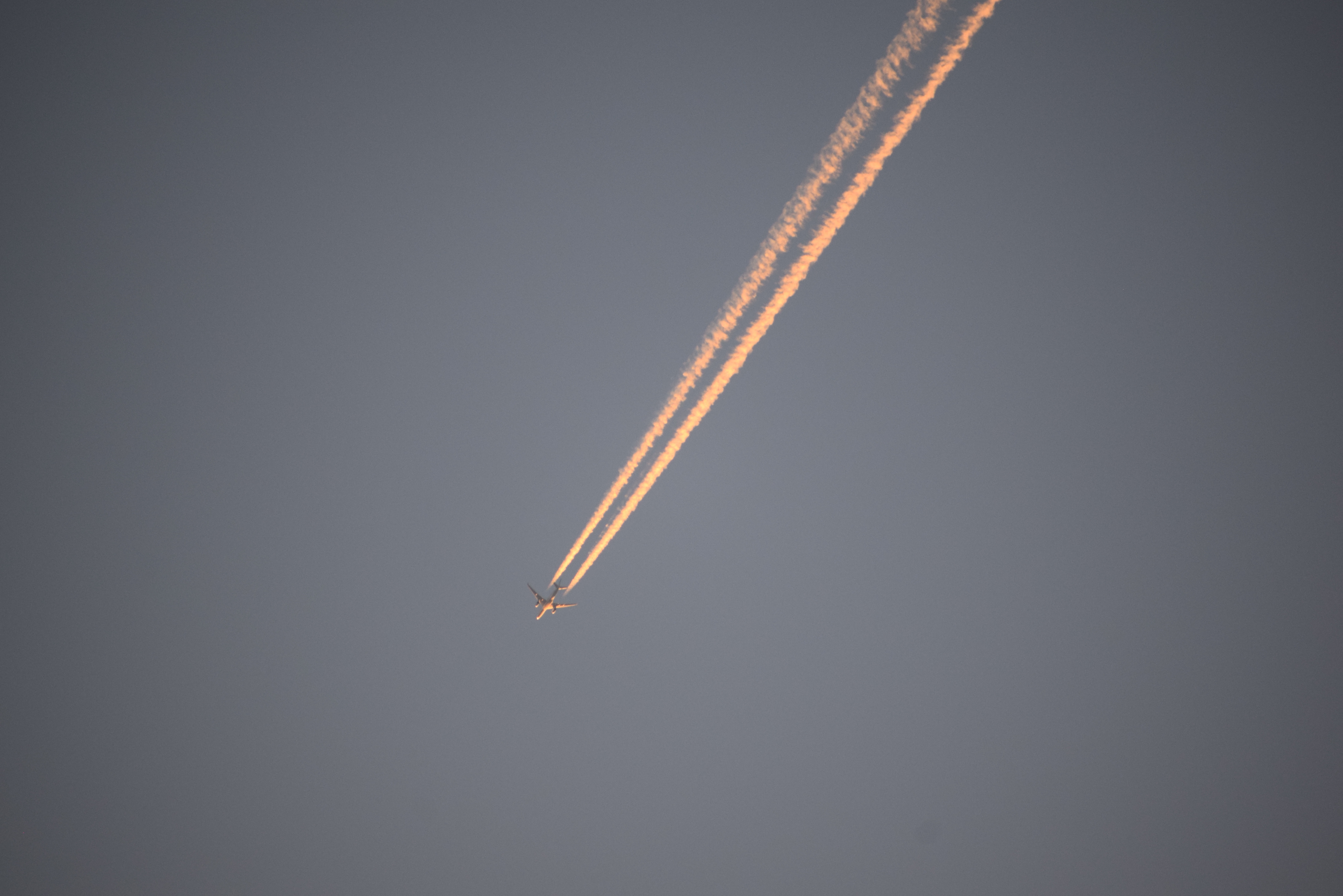A passenger plane high in a clear blue sky as seen from below, with its twin contrails extending out of frame and golden in color in the sunlight.