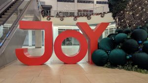 A large, red three-dimensional sign spelling "JOY" is displayed prominently in an indoor space. In the background, there are decorative holiday elements, including garlands and twinkling lights, contributing to a festive christmas atmosphere.