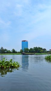 A tranquil scene of a boat gliding on the calm Titas River, Nabinagar, Brahmanbaria, with lush green water plants in the foreground.