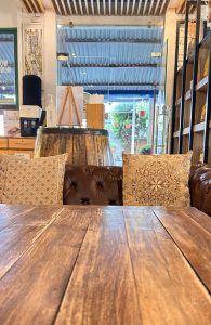 A wooden table with a smooth surface is in the foreground, showcasing rich grains. In the background, a cozy seating area features two decorative pillows with intricate designs resting on a brown leather sofa.
