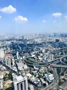 Aerial view of Bangkok city showing dense urban buildings, highways, and flyovers stretching across the city under a clear blue sky.