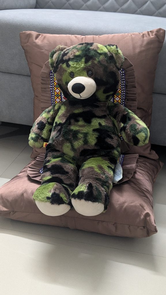 A plush bear with a camouflage pattern in green and black sits on a brown cushion, adorned with decorative elements in the background.