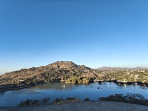 
A scenic view of Mount Abu with rocky mountains, a calm blue lake dotted with boats, and small settlements among green trees under a clear sky.