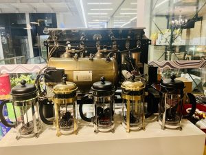 Five French press coffee makers are displayed in a row in front of the exposed brass boiler tank of a coffee machine.