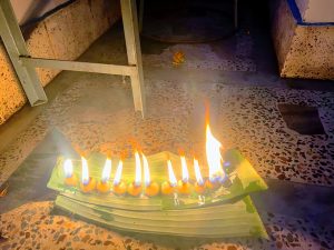 A row of bright cotton oil lamps sits burning on a stack of banana leaves on a tiled floor under a metal frame.