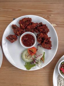 A white plate contains several pieces of dark red fried chicken or meat, a small white bowl of dark dipping sauce, and a garnish of fresh vegetables, all on a light wooden table.
