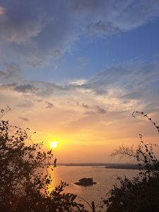 Sunset over a lake. Golden sunlight spills across a tranquil lake at sunset, framed by silhouetted branches and soft clouds drifting through a fading blue-and-amber sky.