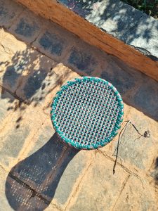 Looking straight down at the top of a stool with a broad woven seat. It's standing on a brick patio. The sun is coming in from the side, showing us via shadow the shape of the lower part. The chair type is called a Muda, often from Punjab.