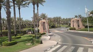 The image shows a landscaped entrance road lined with tall palm trees and neatly trimmed greenery. Two decorative, sand-colored gate structures stand on either side of the roadway, lending the area a resort-style ambiance with a clear blue sky.