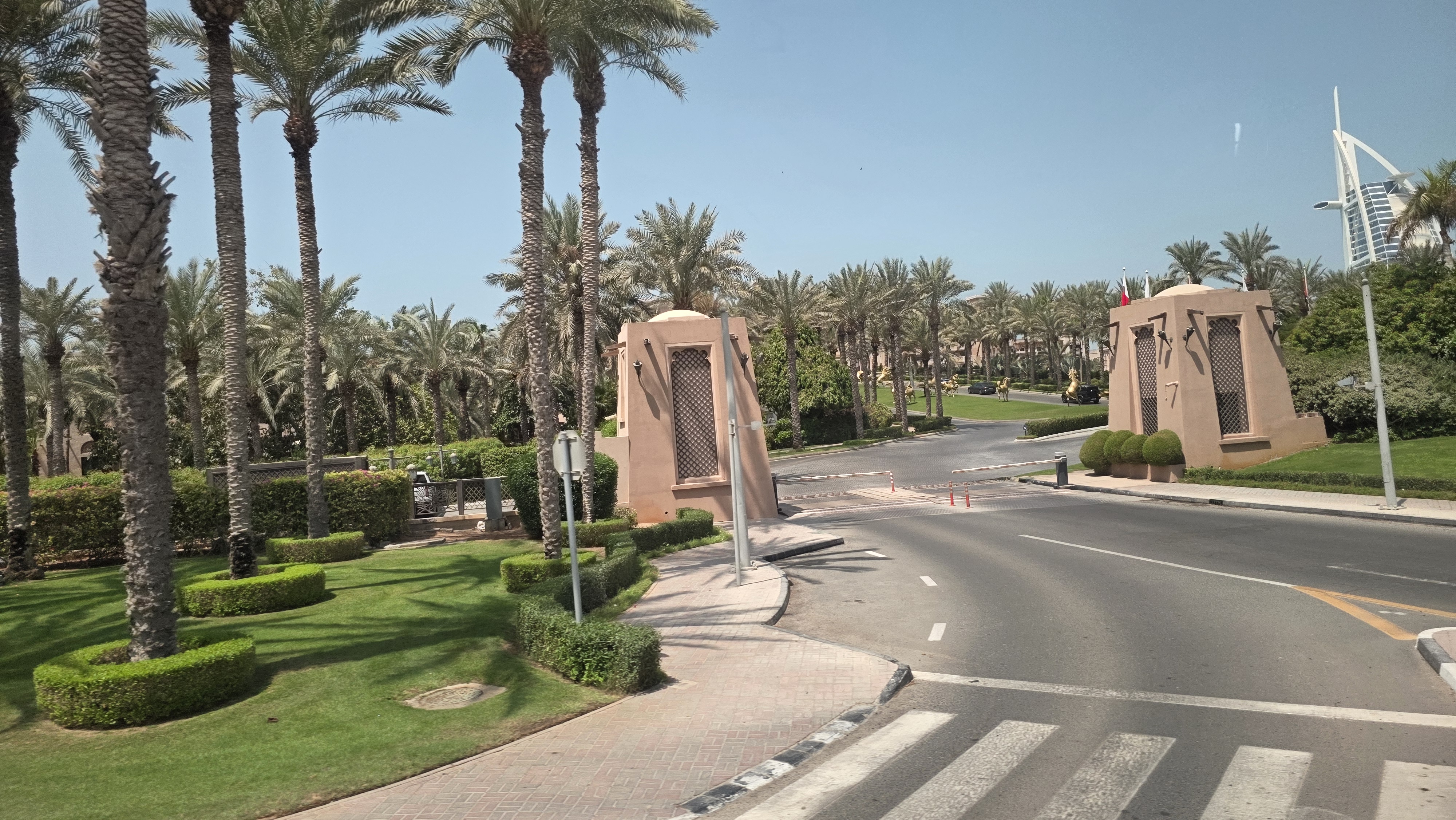 The image shows a landscaped entrance road lined with tall palm trees and neatly trimmed greenery. Two decorative, sand-colored gate structures stand on either side of the roadway, lending the area a resort-style ambiance with a clear blue sky.
