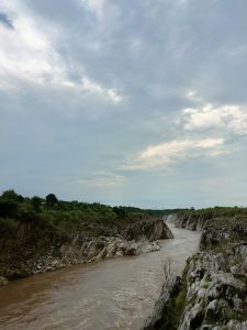 An image of a muddy river flowing through rocky cliffs. The rocky cliff contains green shrubs and vegetation.