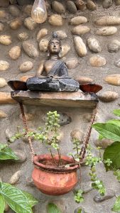 A small Buddha statue sits cross-legged on a wooden shelf against a textured wall made of rounded stones.