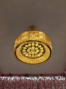 A round, gold-toned crystal chandelier suspended from the ceiling. It emits a warm light through rows of vertical crystals around the side and a pattern of circular crystal elements on the bottom.