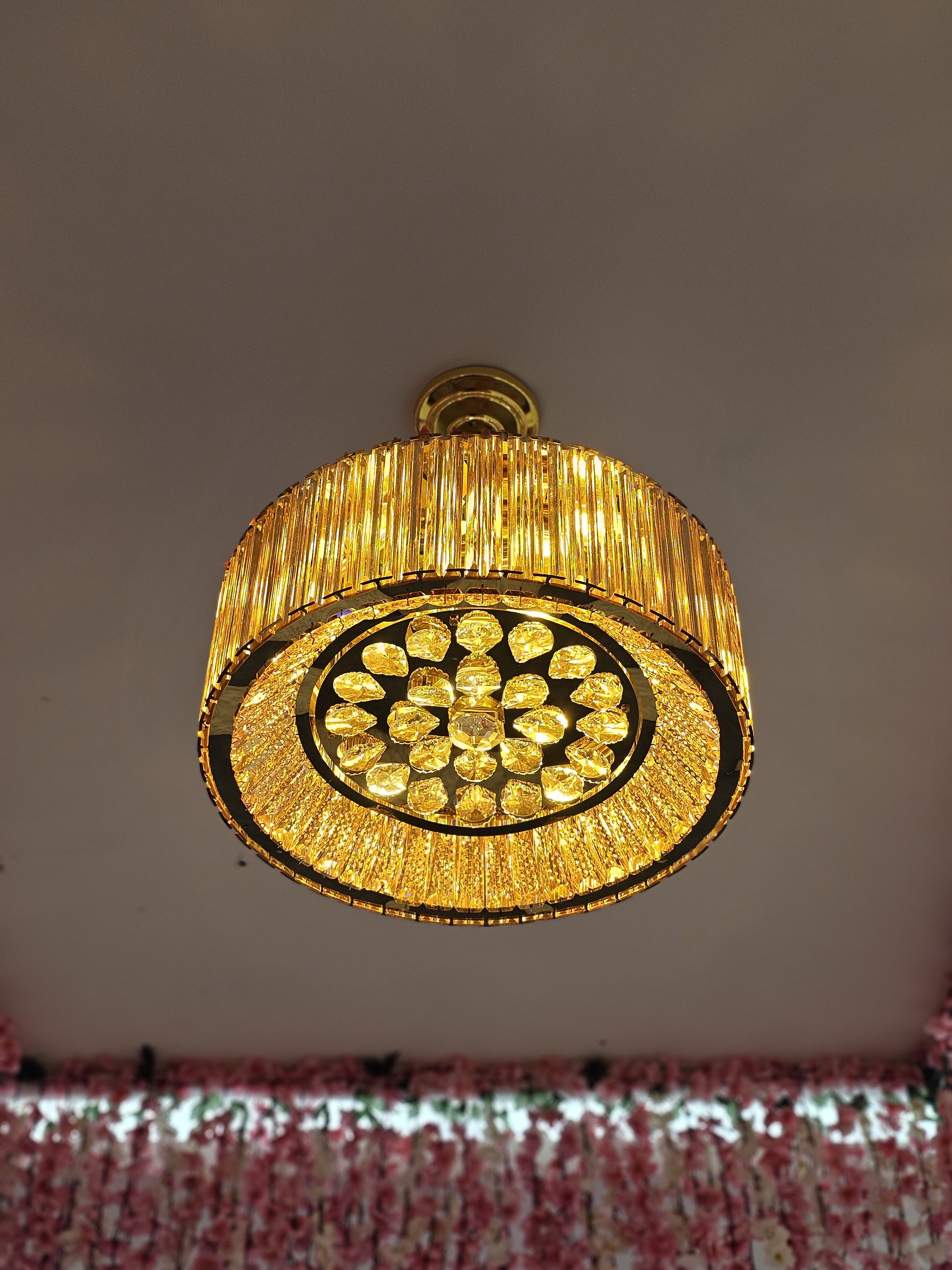 A round, gold-toned crystal chandelier suspended from the ceiling. It emits a warm light through rows of vertical crystals around the side and a pattern of circular crystal elements on the bottom.