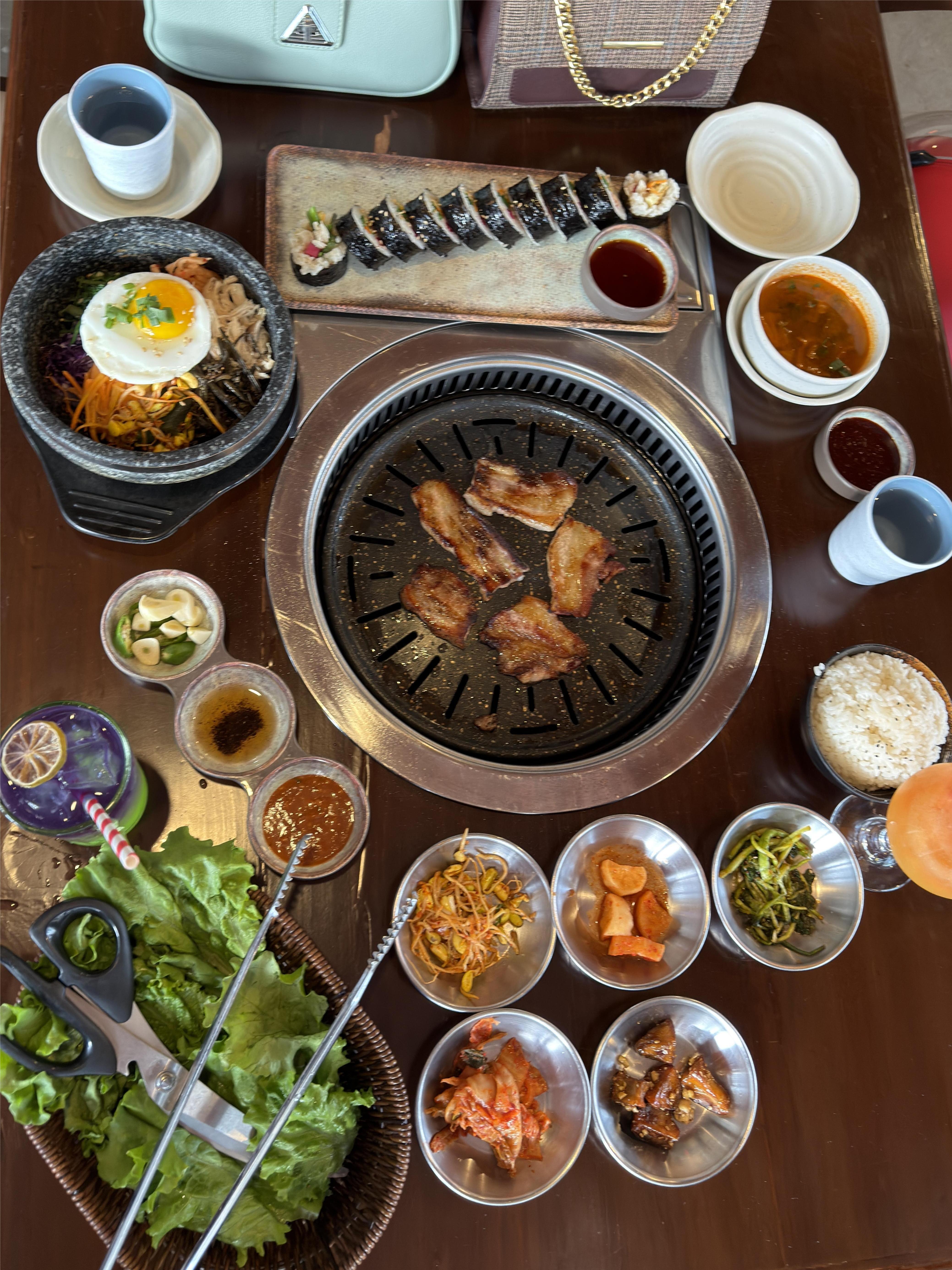 A vibrant spread of Korean cuisine presented on a wooden table. In the center, there's a circular grill with slices of grilled meat. 