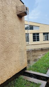 Water flows from a drainpipe on a beige wall into a muddy puddle, with grass and a building in the background.