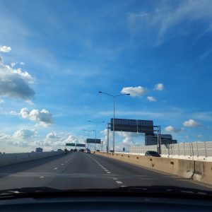 A highway in Thailand, taken from a car, with clear lanes, streetlights, road signs, and buildings in the distance under a bright blue sky.