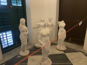 Four white statues of women in draped dresses are displayed indoors, one headless. A red barrier cord crosses diagonally in the foreground.
