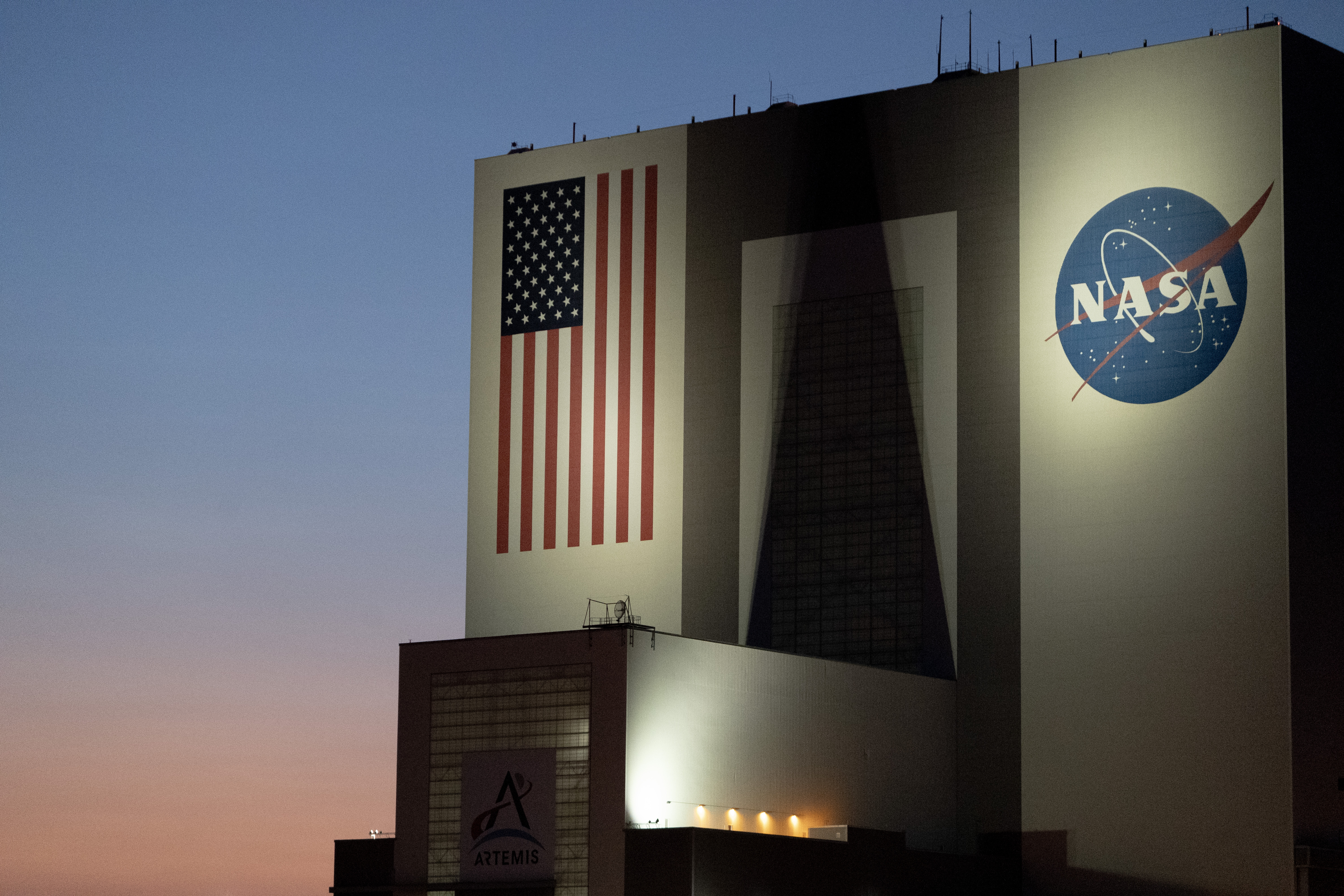 A large white building known as the Vehicle Assembly Building at NASA&rsquo;s Kennedy Space Center, comparable in height to a 50-story structure, set against a gradient sunset sky in the background.