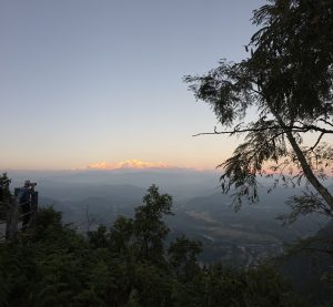 A person stands on a viewing platform, photographing a stunning landscape at sunrise. In the background, snow-capped mountains catch the early light, glowing warmly against a clear sky.