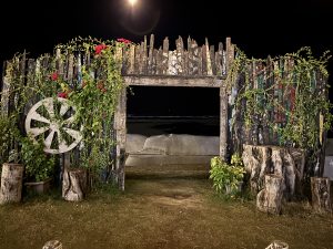 Rustic wooden gate covered with climbing vines and flowers, opening toward a dark beach at night.