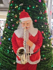 A festive Santa Claus figure playing a shiny saxophone is placed in front of a decorated Christmas tree with glowing lights. This cheerful indoor decoration was captured in Kozhikode, Kerala, bringing holiday joy and musical charm. 