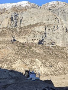 A snow-capped mountain range rises sharply in the background, showcasing rugged cliffs and rocky terrain. In the foreground, a small, white structure with colorful flags stands on a sloped hillside, surrounded by a sparse landscape of brown and green vegetation. 