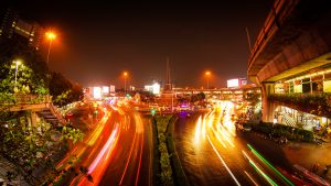 Long exposure photograph of heavy night traffic on a multi-lane highway or main road in a city, showing bright, streaked light trails from car headlights and taillights with elevated infrastructure and city lights in the background.