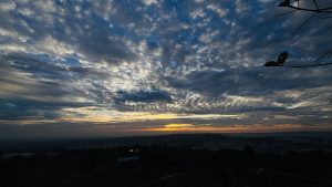 A wide view of the sky filled with thick, textured clouds at sunset. The clouds are dark blue and gray with patches of light. Near the horizon, a warm orange-yellow glow appears. Below, a distant city and hills are faintly visible in shadow, with a few tree branches framing the scene.