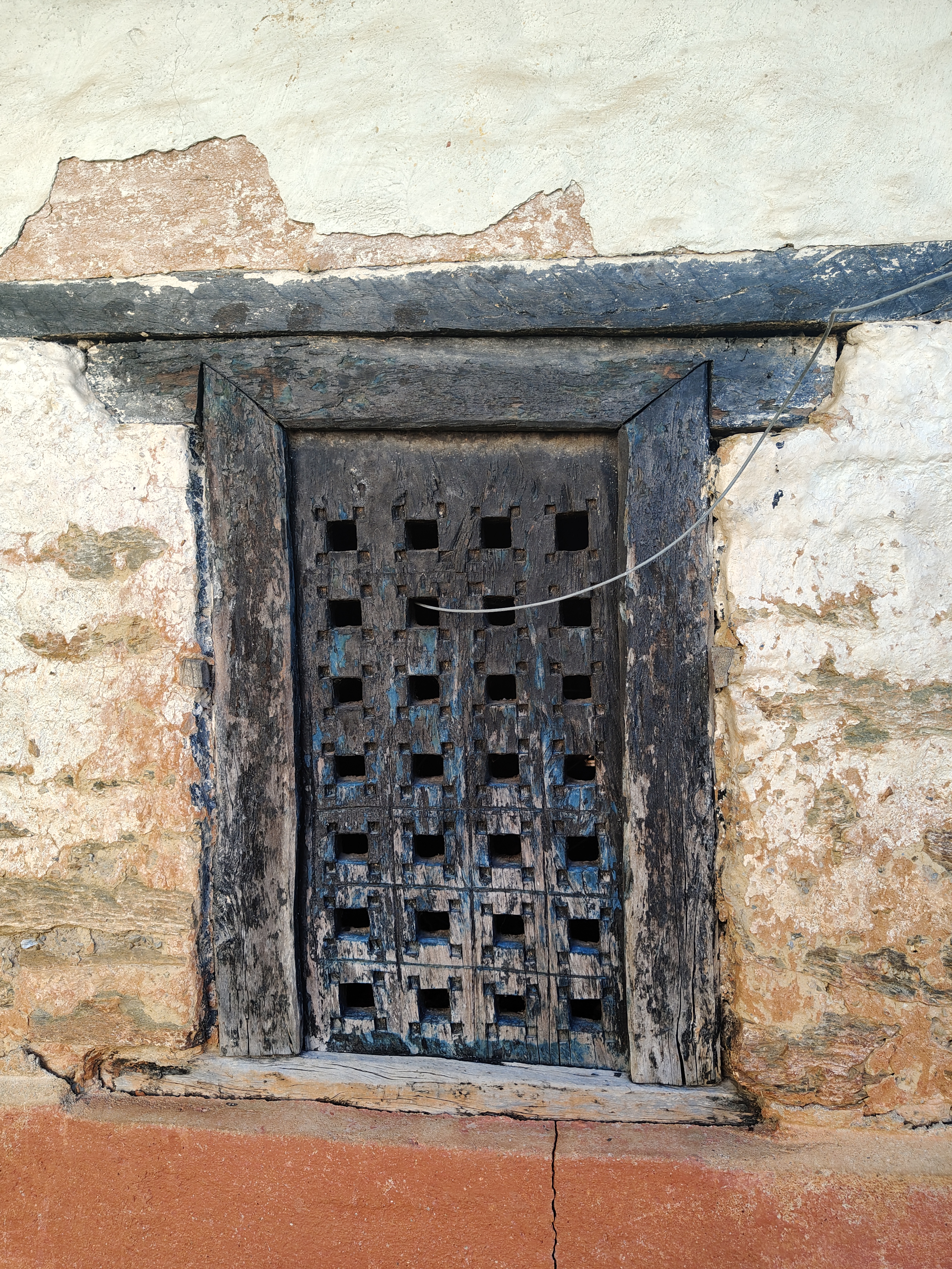 An old wooden window with square cutouts is set in a weathered wall, showing signs of peeling paint and aging. 