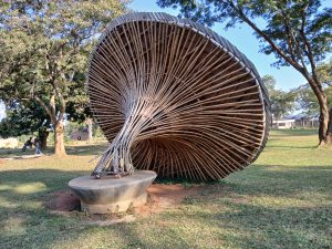 A large, sculptural structure made of bamboo sticks arranged in a spiral pattern, resembling a giant mushroom or a conical hat, is situated in a grassy park area. 