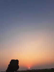 A serene dusk captures the sun glowing at the horizon while a faint crescent moon hangs above, forming a quiet sun-and-moon duo over the silhouetted landscape.