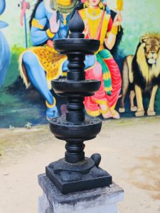 A black stone temple lamp stands in front of a colorful Hindu mural depicting deities. The contrast of stone and paint highlights the spiritual art traditions of Kozhikode, Kerala.