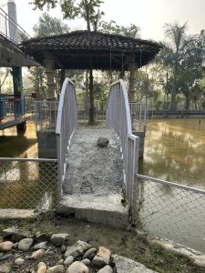 A small, arched footbridge made of concrete and metal leads over to a gazebo with a thatched roof, surrounded by greenery.