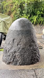 A large, rounded stone resembling an elongated dome sits on a pavement, partially covered in wet sand and grime. 