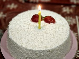 A round white milk cake, covered in shredded coconut, is topped with two fresh strawberries and a single lit yellow candle, sitting on a pink board.