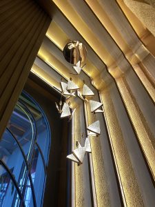 Elegant chandelier with triangular glass pendants hangs against a textured wall. Warm, ambient lighting creates a sophisticated, modern atmosphere.