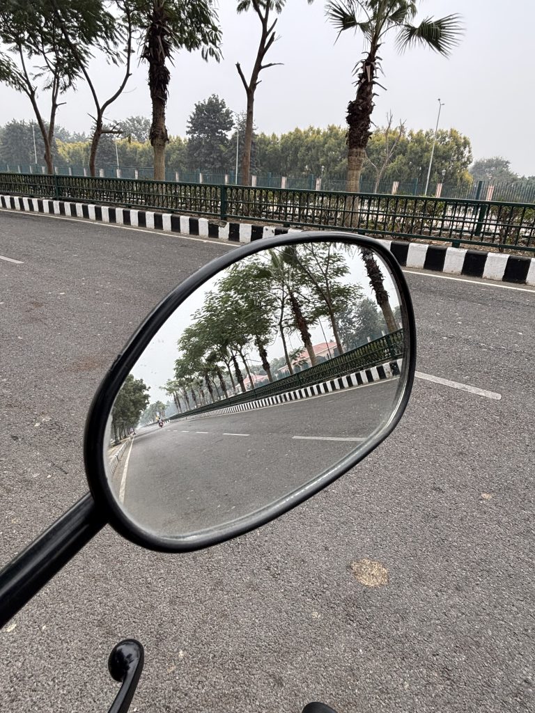 A close-up view of a motorcycle’s side mirror reflecting a deserted road lined with trees and a green fence. The road is largely empty, with a faint silhouette of a motorcycle visible in the distance.