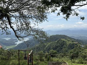 A scenic view from a high point, featuring a lush green landscape with rolling hills and a lake winding through the valley below. 