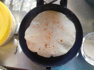 A brownish, round flatbread is cooking in a black frying pan on a metallic surface. 