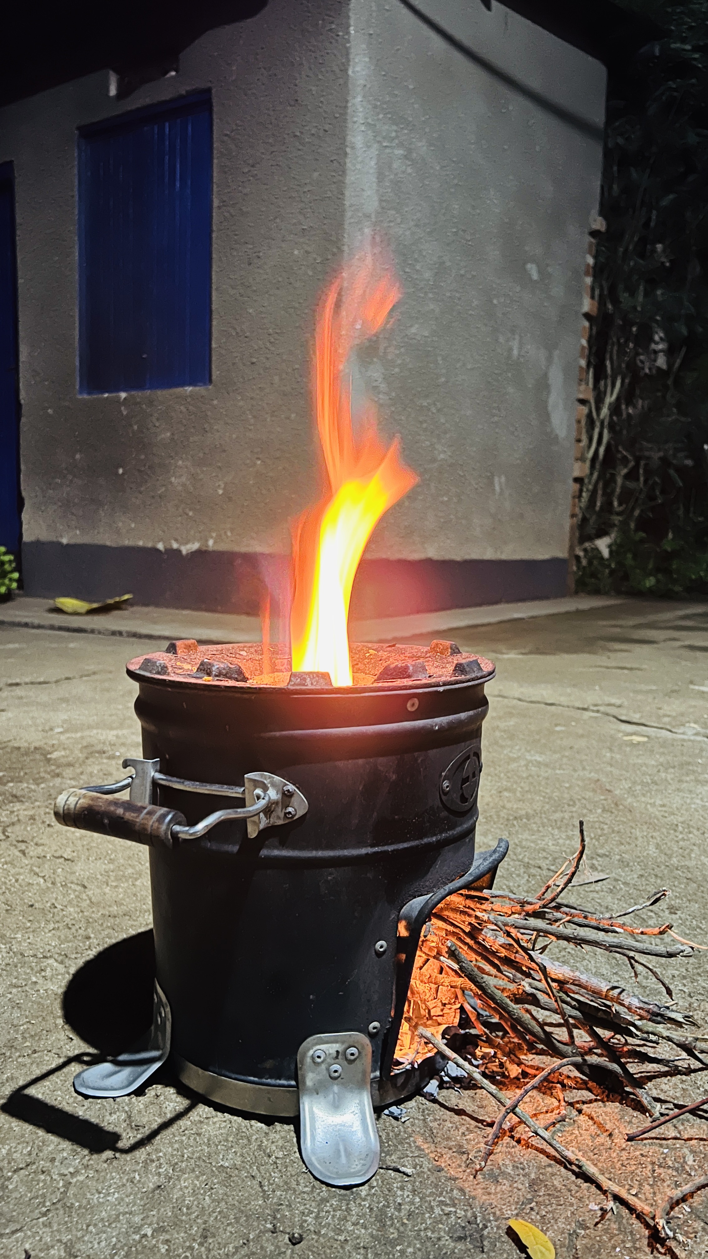 A black metal stove is positioned on a concrete surface, with flames and glowing embers visible at the top and side. 