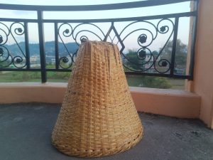 A woven, conical basket made of natural fibers stands on a concrete surface, with a decorative black railing in the background. 