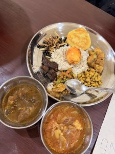 A silver plate filled with various dishes, including a mound of oats at the center, topped with a golden-brown egg. Surrounding the oats are pieces of fried food, vegetables, and small black beans