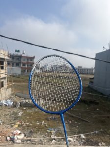 A close-up of a blue badminton racket held up against a backdrop of a cloudy sky and a construction site.
