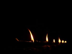 A row of small oil lamps glows gently in the dark, creating a calm and peaceful scene. The soft flames stand out against the black background. Captured at Velliparamba, Kozhikode, this photo reflects quiet evening light and simplicity.