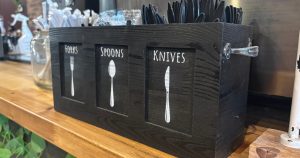 A black wooden box with three sections labelled "Forks", "Spoons", and "Knives". There's a picture of each item on its section, and each section is filled with plastic utensils. 