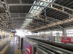Wide-angle view of a modern metro station with steel beams, skylights, and empty platforms. Bright and spacious with signs and digital displays overhead.